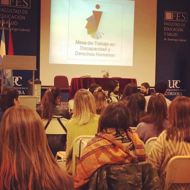 Foto tomada en una charla. Se ve gente de atrás sentada mirando hacia una pantalla donde se proyecta el logo de la mesa. También se ven carteles de la UPC.