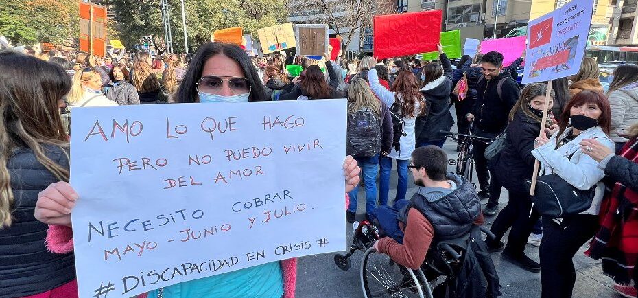 Foto de la movilización de esta mañana frente al Patio Olmos. Se ve un grupo de personas manifestando, en primer plano una mujer sostiene cartel que dice: "Amo lo que hago pero no puedo vivir del amor. Necesito cobrar mayo, junio y julio. #DiscapacidadenCrisis." Fuente periodista Andrés Ferreyra
